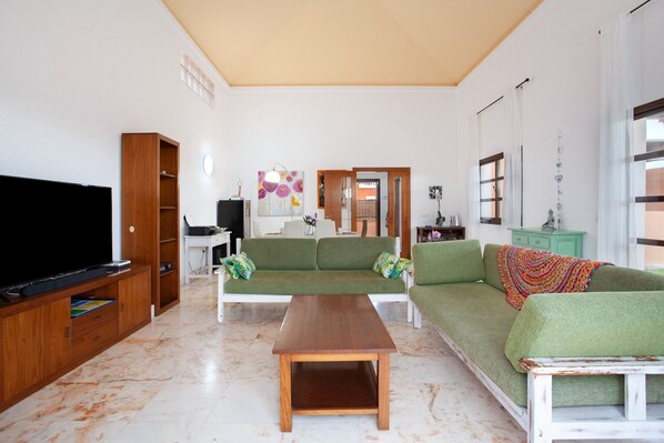 A living room with a flat-screen TV, a wooden entertainment unit, a green sofa, a white coffee table, and a dining area with a table and chairs.