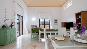 A living room with a dining area, a green sofa, white chairs, a television, and a cabinet.