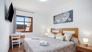 A bedroom with a bed, a desk, a chair, a TV, and a painting on the wall.