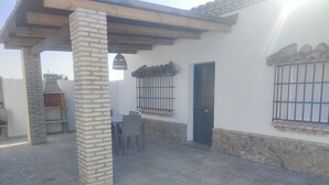 Een terras met een bakstenen pilaar, een barbecue, een tafel met stoelen en een deur met tralies.