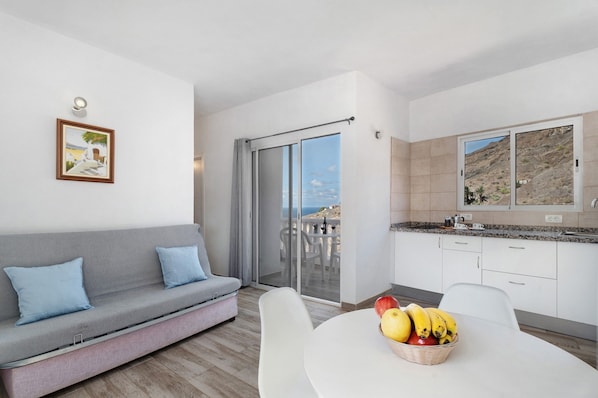 A modern living room with a sofa, a dining table with fruit, a kitchen with a large window, and a balcony with a view.