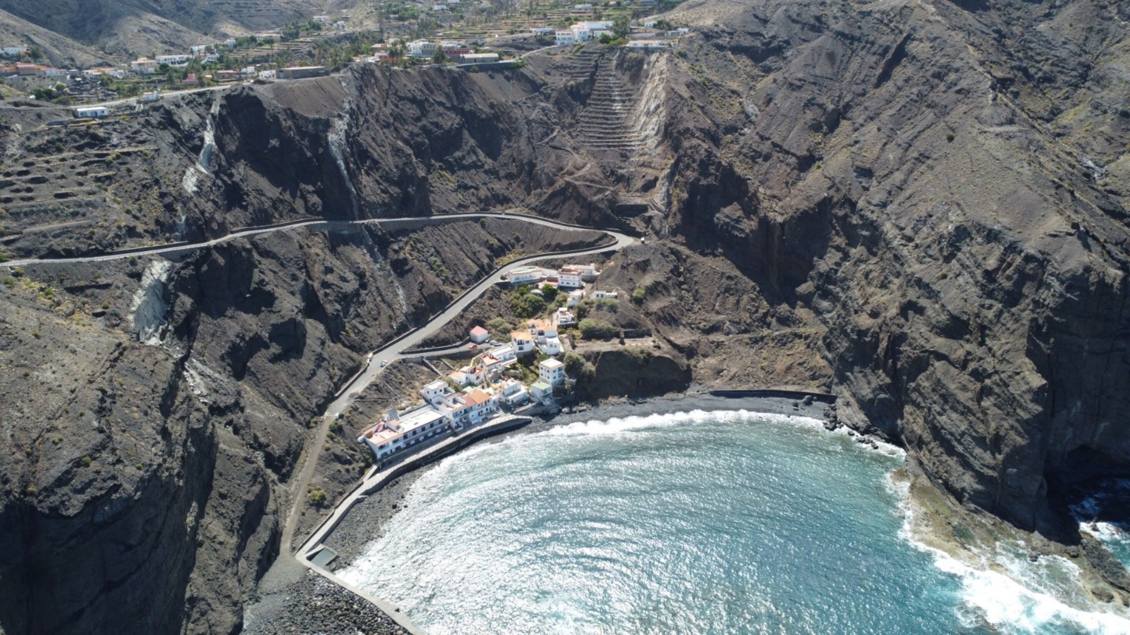 Zona costera con una carretera que conduce a un poblado, acantilados rocosos y una masa de agua.