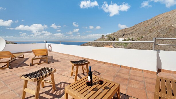 A terrace with wooden furniture, a bottle of wine, and a view of the sea and hills.
