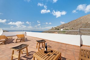 Une terrasse meublée de mobilier en bois, une bouteille de vin et une vue sur la mer et les collines.