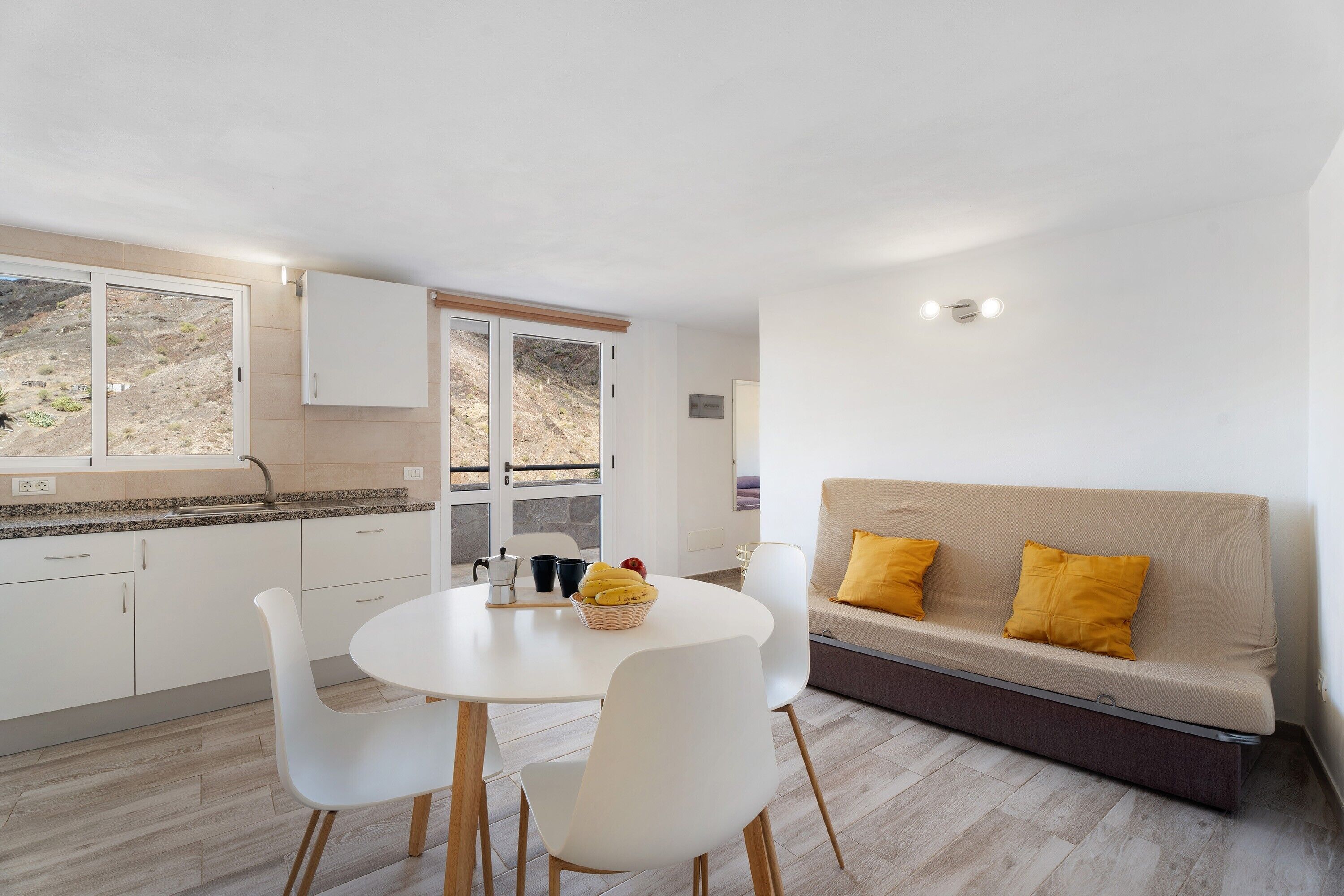 Una cocina moderna con armarios blancos, una mesa de comedor con sillas, un sofá con cojines amarillos y vistas a un paisaje montañoso a través de la ventana.