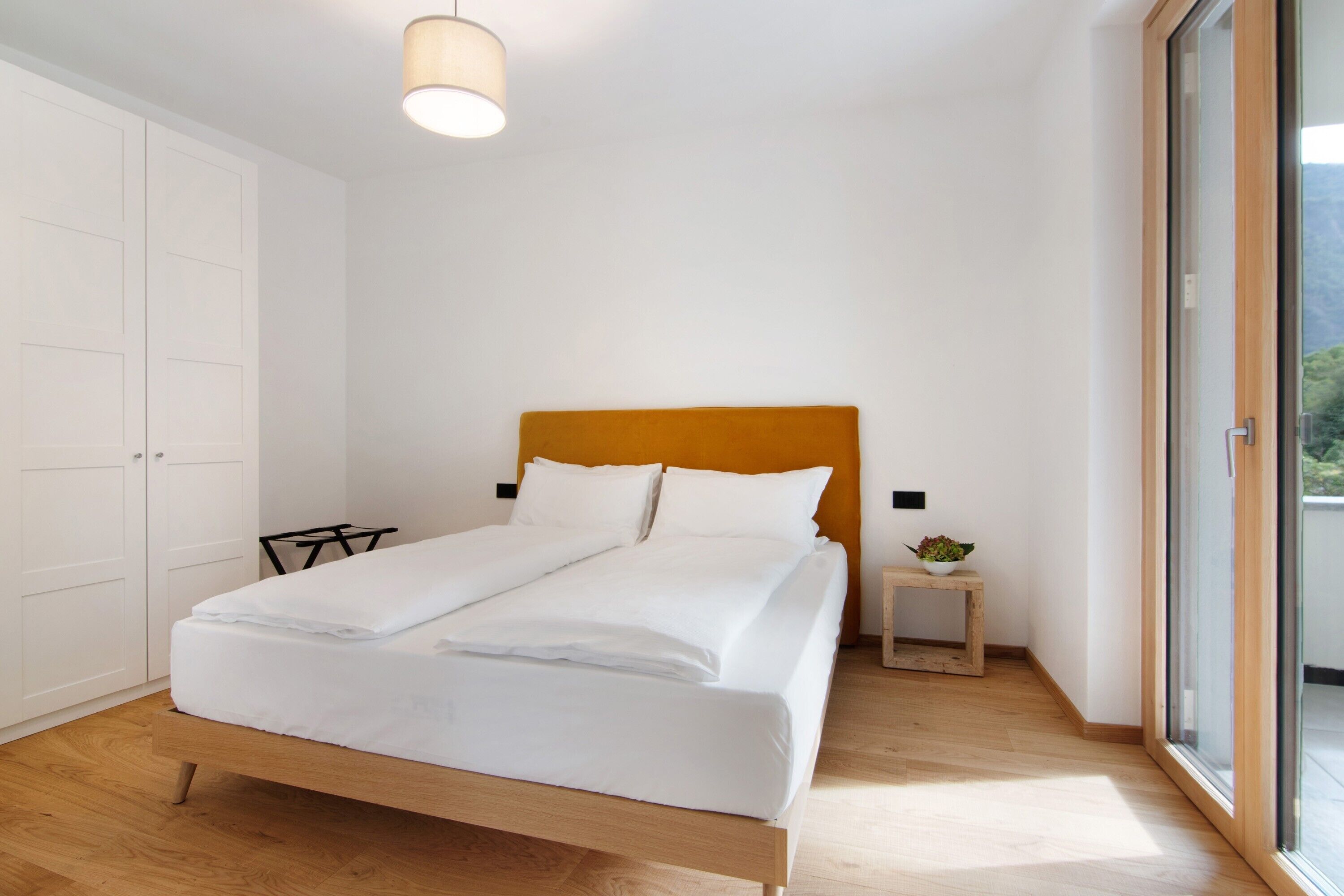 Un dormitorio con cama, una mesita de noche con planta, un armario y una puerta corrediza de vidrio al exterior.