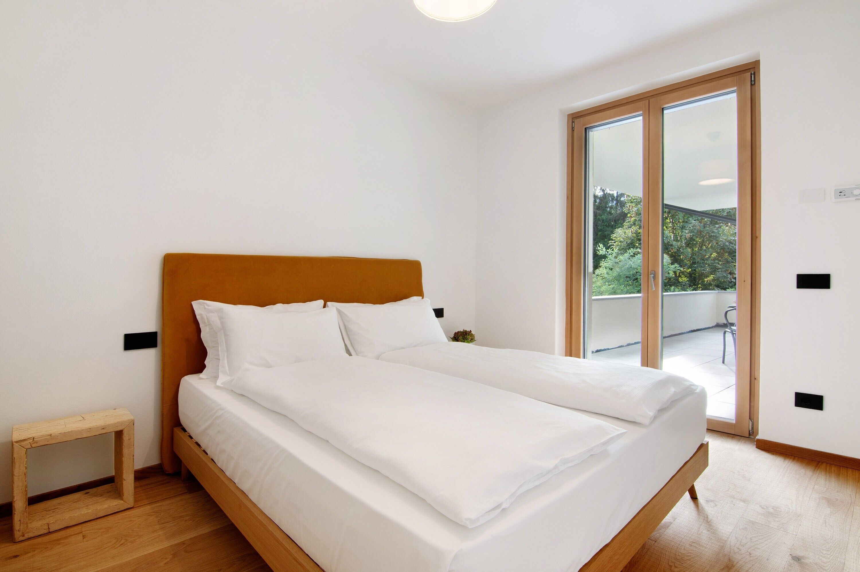 Un dormitorio con una cama de madera, ropa de cama blanca, un taburete de madera pequeño y un ventanal corredero que da a un balcón exterior.