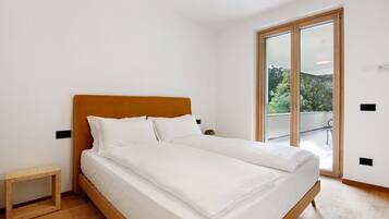 Un dormitorio con una cama de madera, ropa de cama blanca, un taburete de madera pequeño y un ventanal corredero que da a un balcón exterior.