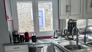 A modern kitchen with white cabinets, a stainless steel dishwasher, a double sink, and a window with a view of a stone wall and blue door. - Holiday Home 'Chez Brigitte' with Private Terrace, Wi-Fi and Air Conditioning (Chatillon sur marne)