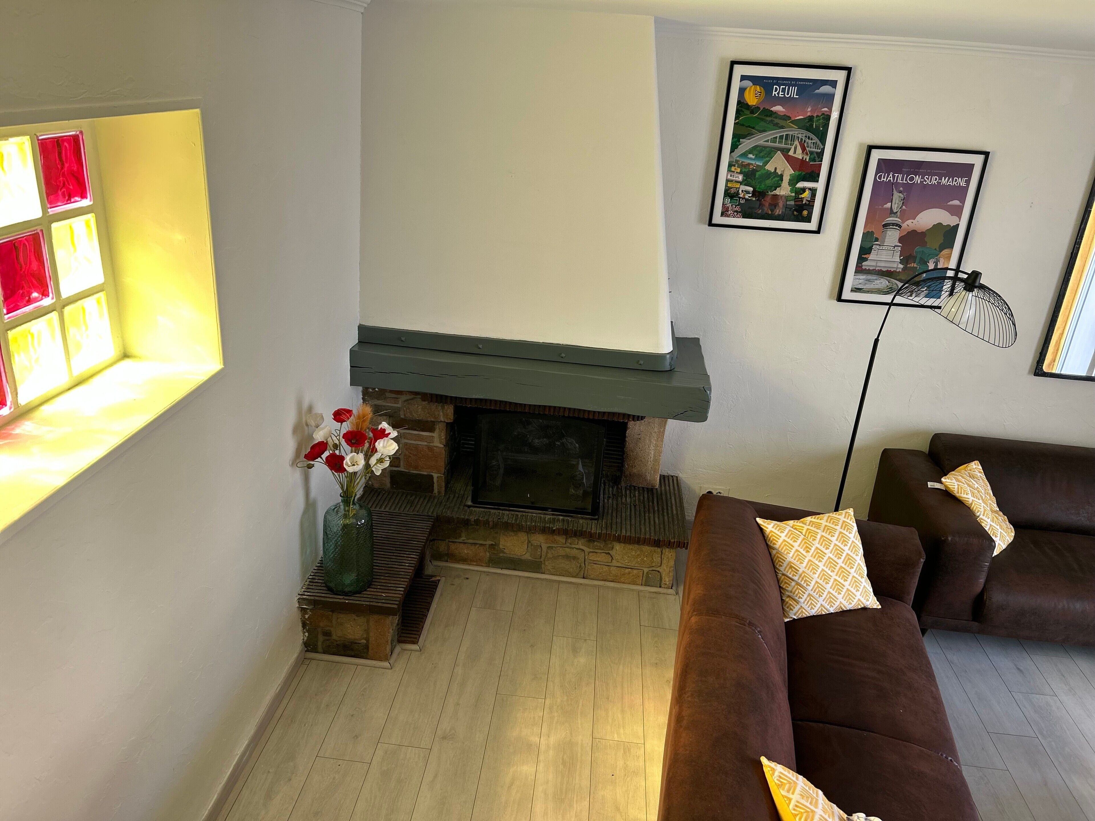 Un salon avec une cheminée, un canapé marron, un lampadaire et deux tableaux encadrés accrochés au mur.