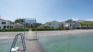 Een houten terras dat leidt naar een helder zwembad met een metalen trap, omgeven door woongebouwen en een strand.