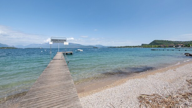 Een houten steiger die zich uitstrekt over een kalm meer met helder water, omgeven door een kiezelstrand en verre bergen.