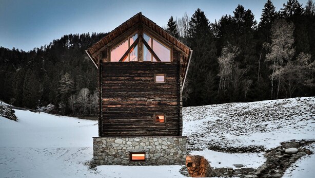 Uma cabana de madeira com base de pedra, rodeada por neve e árvores.