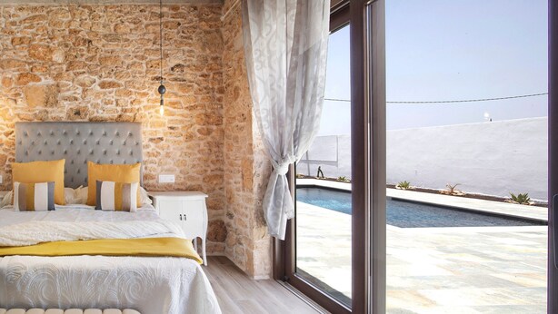 A bedroom with a stone wall, a bed with a tufted headboard, a bedside table, and a view of a swimming pool. - Casa Rural Berrenda with Sea Views, Private Terrace, and Wi-Fi (Tiscamanita)