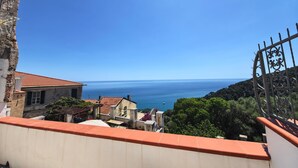 Vista sulla costa con cielo sereno, un balcone con tetto in tegole rosse e una veduta distante del mare.