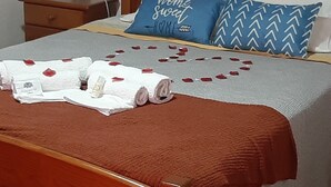 Un lit bien fait, avec une tête de lit marron, un oreiller bleu orné du texte « chez soi, on est bien », une table de chevet en bois avec une lampe, et une composition de pétales de rose rouge sur le lit.