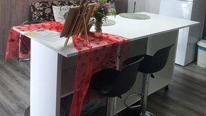 Une cuisine moderne avec un plan de travail blanc, deux tabourets de bar noirs, une petite table recouverte d’une nappe rouge et un réfrigérateur.