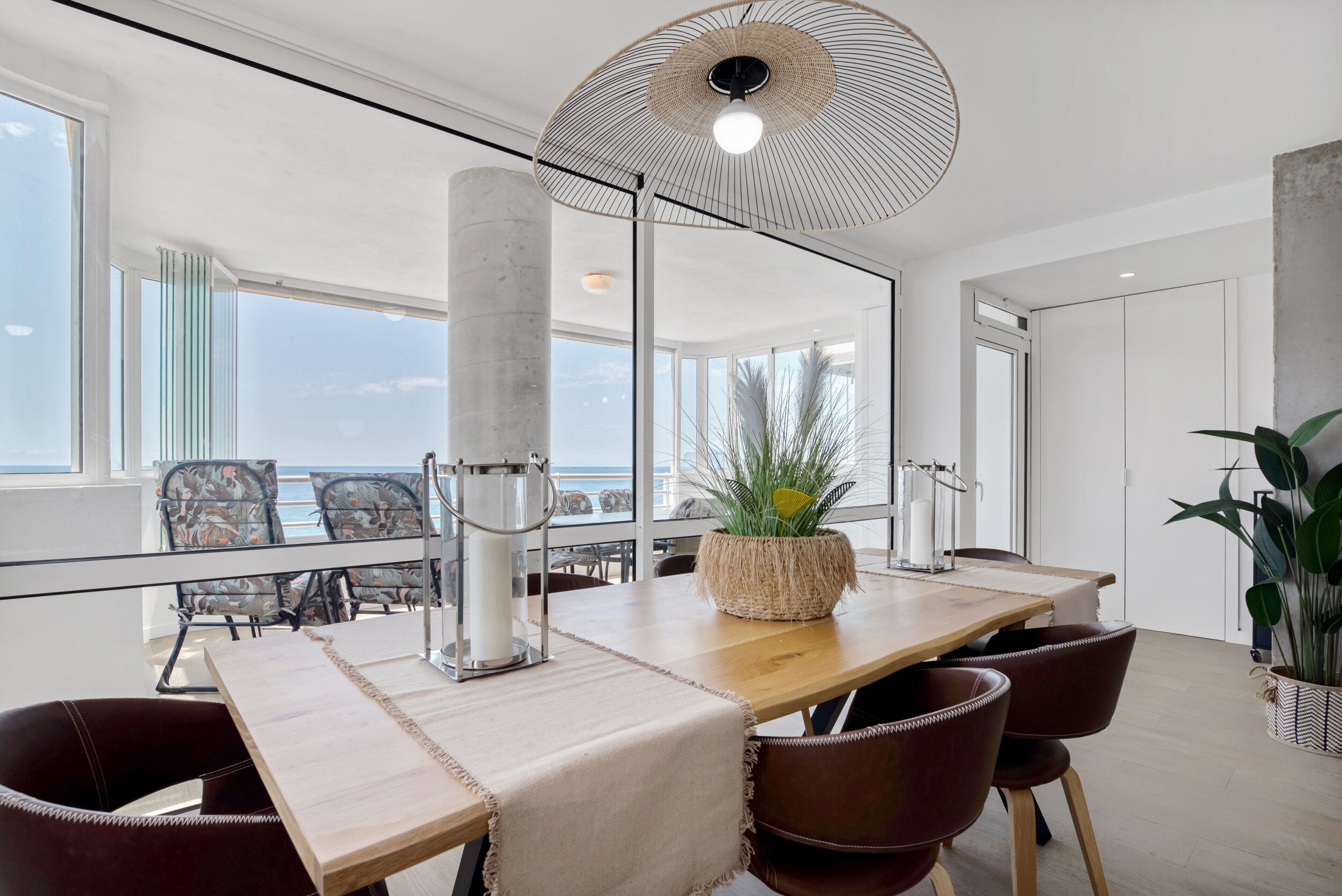 A modern dining area with a wooden table, leather chairs, a hanging light fixture, and a view of the ocean.