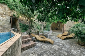 A stone building with a swimming pool, wooden loungers, and a table with chairs.
