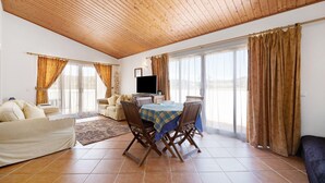 Uma sala de estar ampla com teto de madeira, uma mesa de jantar com cadeiras, um sofá e uma televisão.