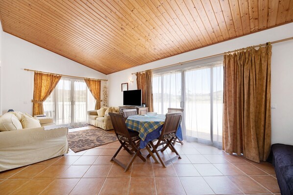A spacious living area with a wooden ceiling, a dining table with chairs, a sofa, and a TV.