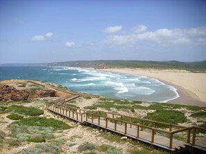 Un paysage côtier avec une promenade en bois menant à une plage, le tout entouré d’un terrain rocheux et d’une végétation clairsemée.