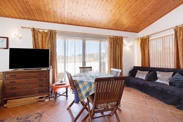 Une pièce avec un plafond en bois, un téléviseur posé sur une commode, une table à manger avec des chaises, un canapé et de grandes fenêtres avec des rideaux.