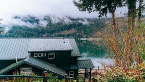 A cabin with a metal roof and multiple windows, situated near a lake with forested mountains in the background. - Emerald Haven Lakeside Paradise, Ultimate Retreat (Port Angeles)
