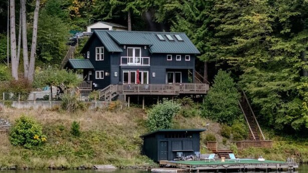 A dark-colored cabin with a deck, situated by a lake surrounded by dense forest. - Emerald Haven Lakeside Paradise, Ultimate Retreat (Port Angeles)