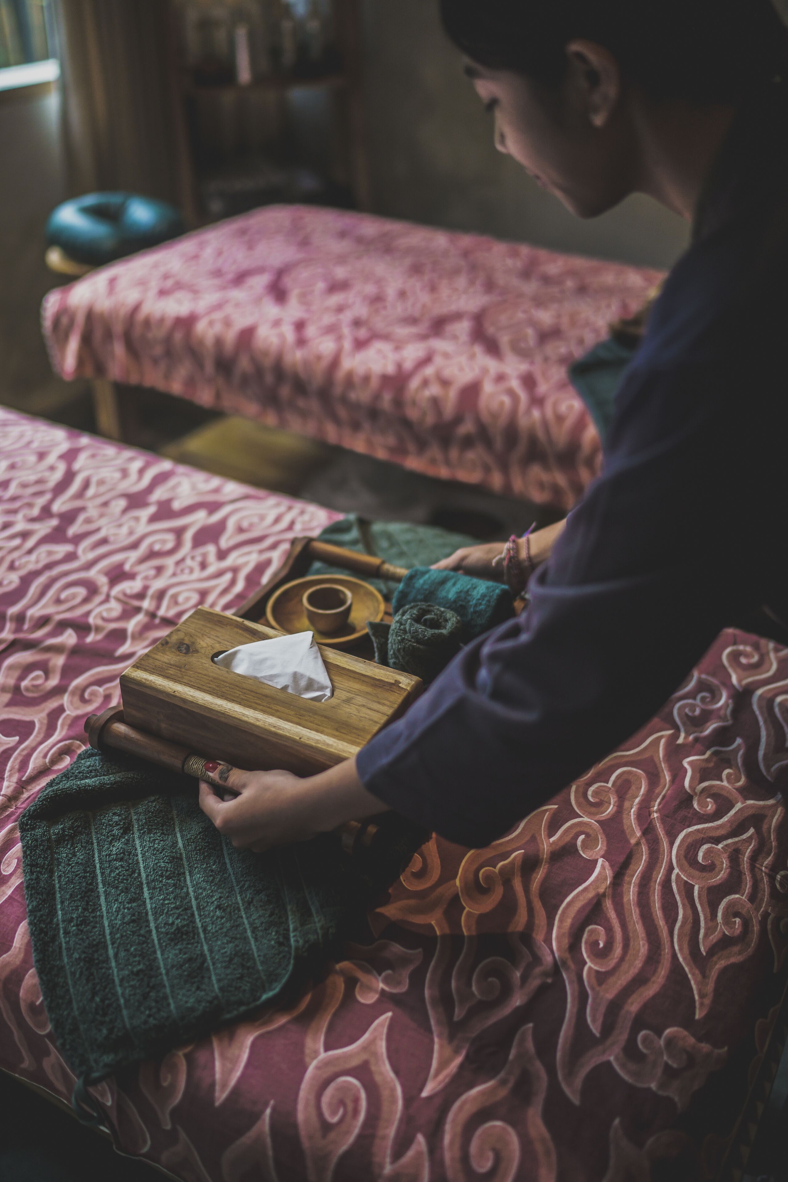 Une personne est assise sur un lit recouvert d’une couvre-lit à motifs, tenant une boîte de mouchoirs et une serviette verte.