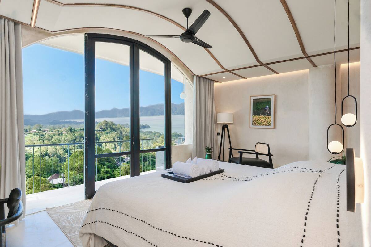 Quarto de hotel com uma cama grande, uma escrivaninha, uma cadeira, um ventilador de teto e vista para uma paisagem montanhosa.