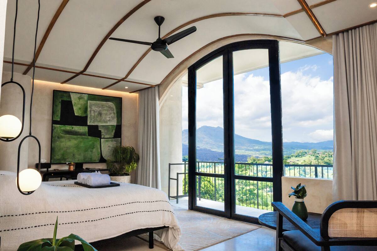 Quarto com uma cama grande, ventilador de teto, vista para o exterior e varanda com guarda-corpo.