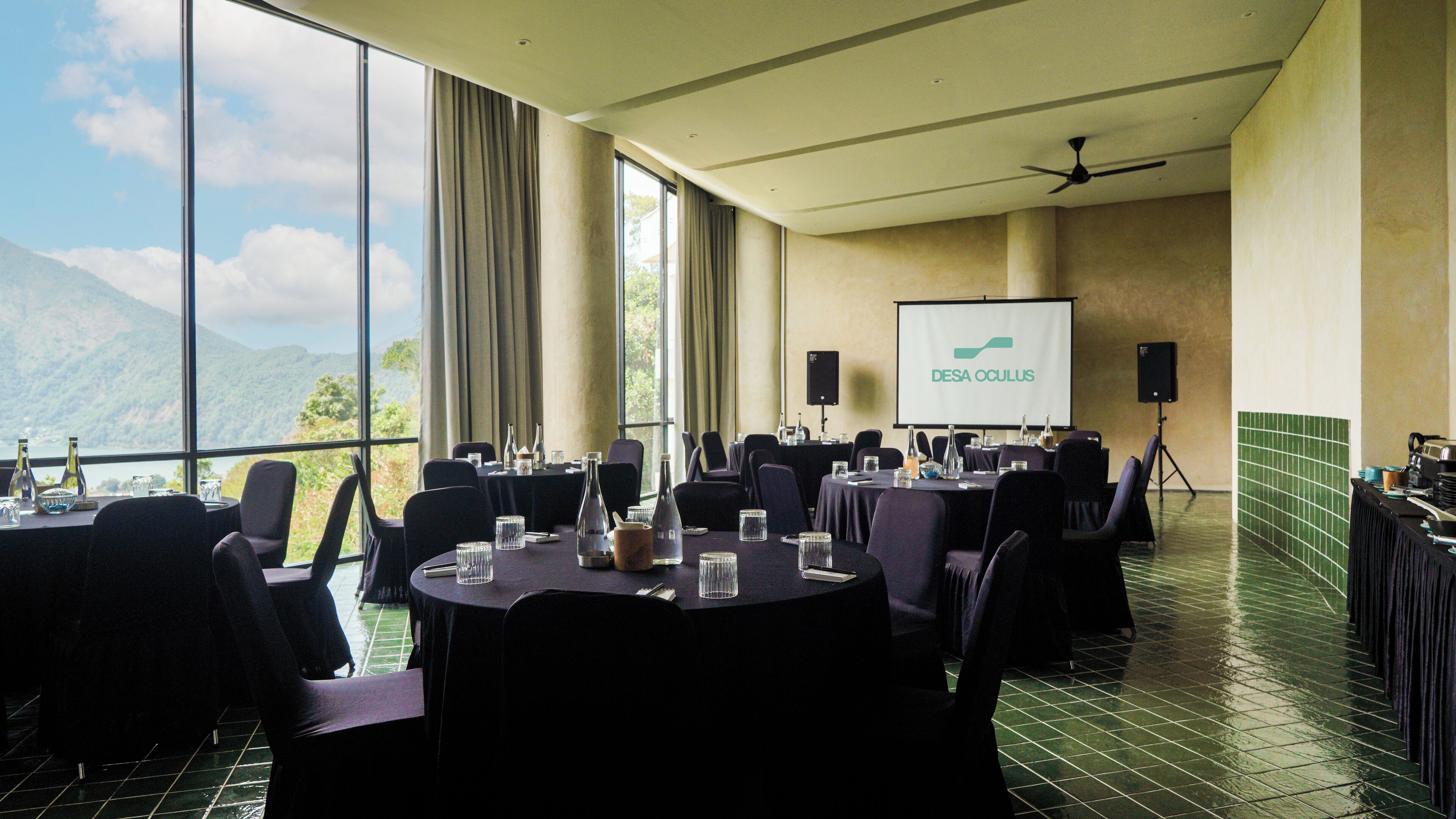 Une salle de conférence avec des tables disposées pour une réunion, un écran de projection affichant le texte « DESA OCULUS », et une vue sur les montagnes à travers de grandes fenêtres.