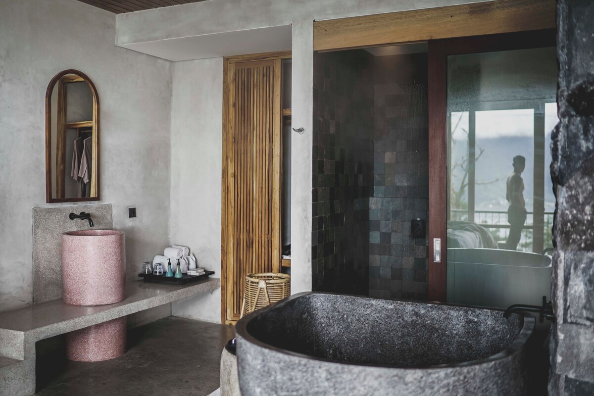 Un baño con una bañera de piedra grande, un lavamanos rosa y una puerta de madera que da a un balcón.