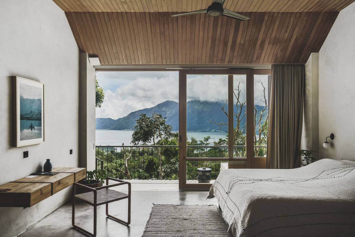 Quarto com uma grande janela, teto de madeira e vista para montanhas e água.