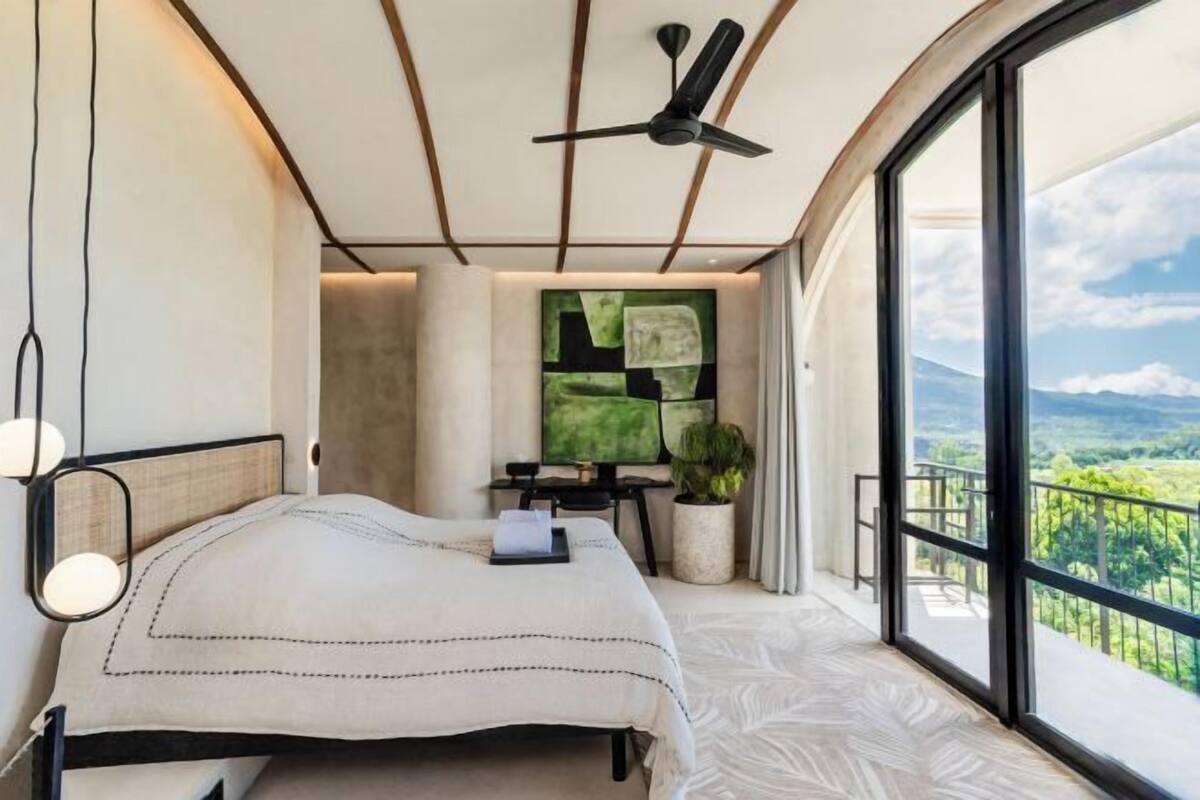 Quarto com cama, ventilador de teto, escrivaninha com computador, janela com vista e planta em vaso.