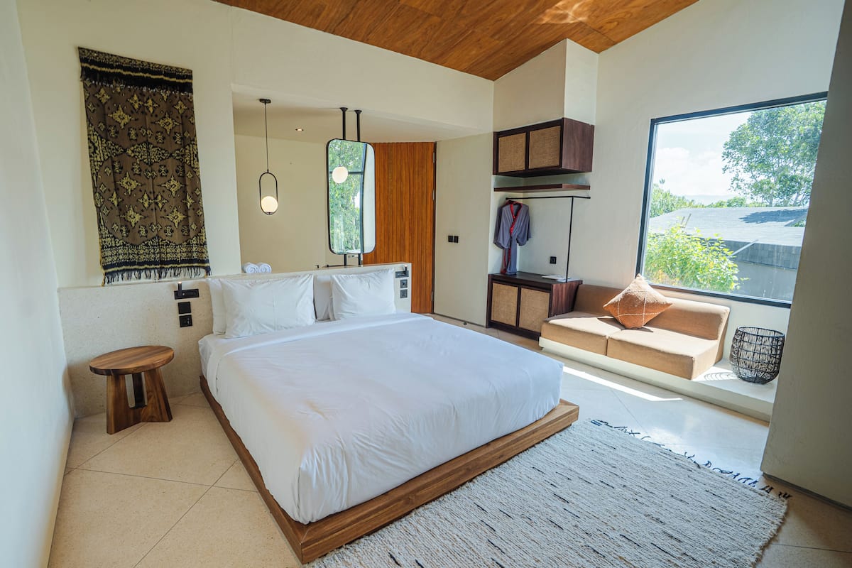 Quarto moderno com cama grande, cabeceira de madeira, um banquinho pequeno, um sofá e uma janela ampla com vista para o verde.