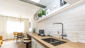 A modern kitchen with a wooden countertop, a sink, and a built-in oven. There are bar stools and a dining area with a table and chairs. - Apartamento Primavera (Parede)