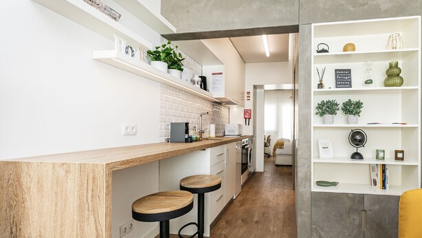A modern kitchen with wooden bar stools, a built-in coffee machine, and a shelf with decorative items. - Apartamento Primavera (Parede)
