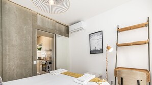 A modern bedroom with a large bed, a desk, a chair, and a kitchenette in the background. - Apartamento Primavera (Parede)