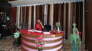 A reception desk with a woman seated behind it, a telephone, a small table with a vase of flowers, and a fan in the background. - Ugte Hotel (Rishikesh)