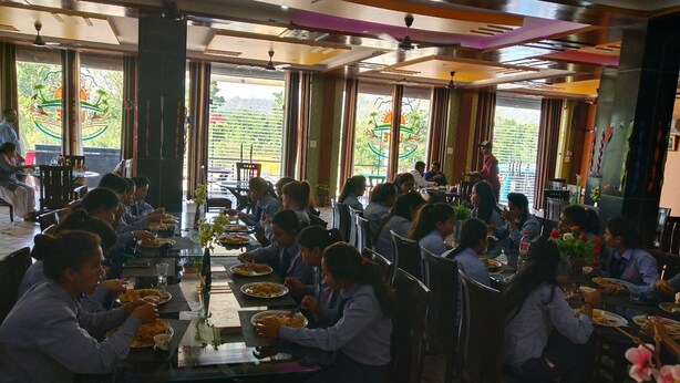 A dining area with tables and chairs, large windows with decorative elements, and ceiling fans. - Ugte Hotel (Rishikesh)