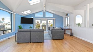 Een ruime woonkamer met een grote grijze modulaire bank, een flatscreen televisie die aan de muur is bevestigd, en een eethoek met een houten tafel en stoelen.