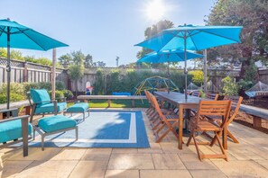 An outdoor patio with a wooden table and chairs, blue umbrellas, and a play structure in the background. - Oceanview Family Playground (Santa Cruz)