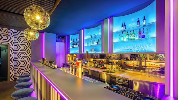 Una zona de bar con diseño moderno, que cuenta con una barra larga, múltiples estantes llenos de botellas y una iluminación particular.