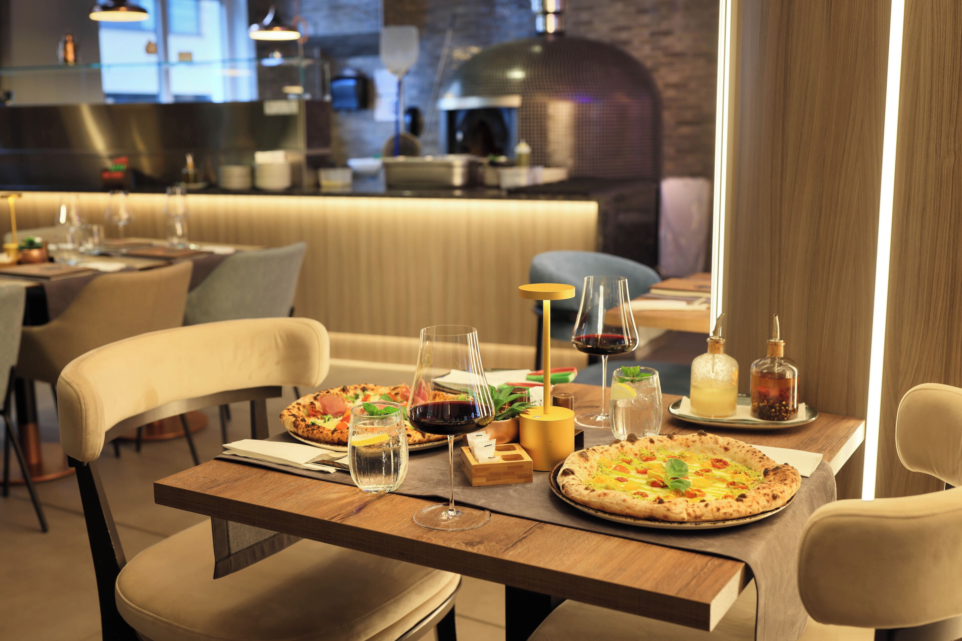 Un restaurante con una mesa de madera dispuesta para dos personas, que presenta pizzas, copas de vino y una vela.