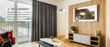 A modern living room with a sofa, a dining table, a bench, a TV mounted on a wooden wall, and a balcony with a view of buildings.