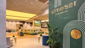Un moderno vestíbulo con una pared verde que presenta el nombre 'somerset' y 'SÖHNBRÜNN'. Hay zonas de estar con sofás y sillas, y una gran planta en maceta al frente.