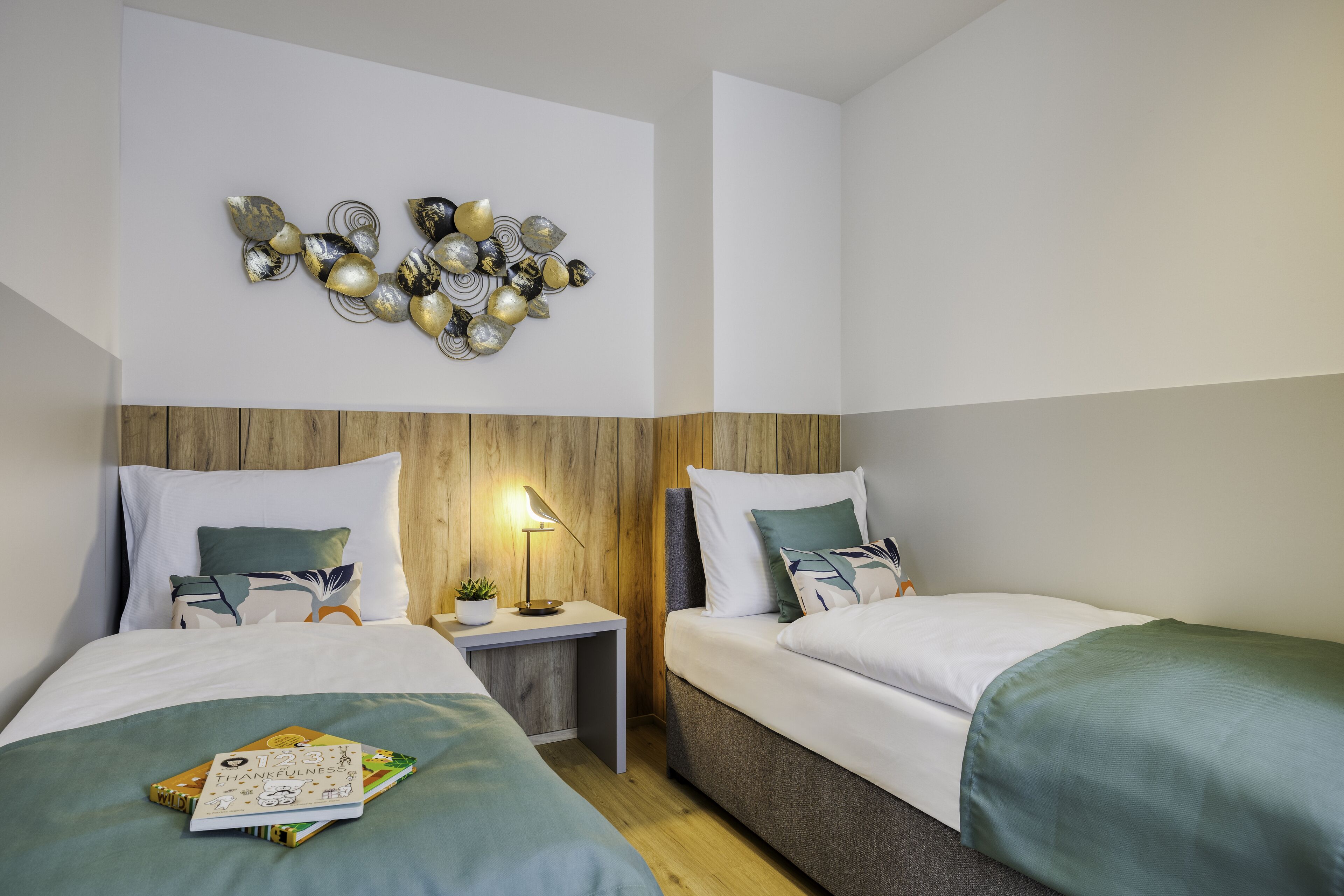A modern hotel room with two beds, a wooden headboard, a wall art piece, and a bedside table with a lamp.