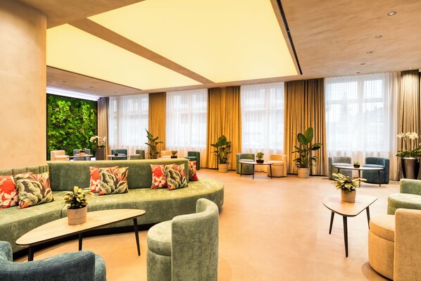 A modern hotel lobby with a curved green sofa, multiple seating areas, and a large window with curtains.